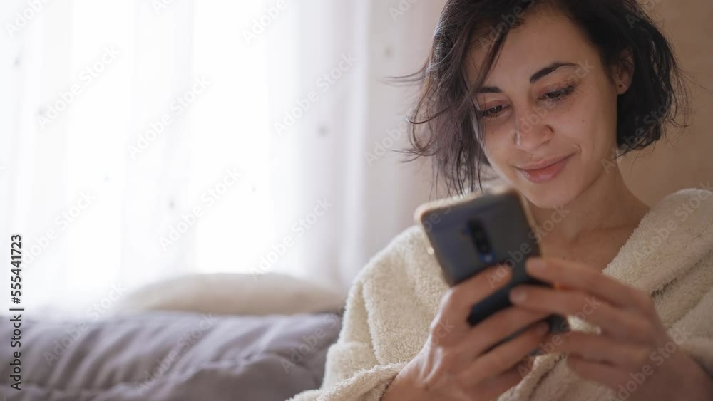 beautiful dreamy brunette woman in warm white bathrobe sitts at cozy home on coach using phone, texting, messaging or looking social networks beautiful dreamy brunette woman in warm white bathrobe sitts at cozy home on coach using phone, texting, messaging or looking social networks