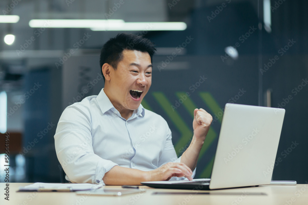 Young man businessman freelancer Asian looks at the laptop monitor in the office, received important good news, rejoices, points with his hand yes, shouts with happiness. Young man businessman freelancer Asian looks at the laptop monitor in the office, received important good news, rejoices, points with his hand yes, shouts with happiness.