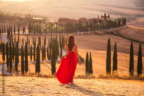 A girl at sunset in a red dress on a field in Italian Tuscany. Val d'Orcia. Beautiful landscape scenery at sunset of Tuscany in Italy