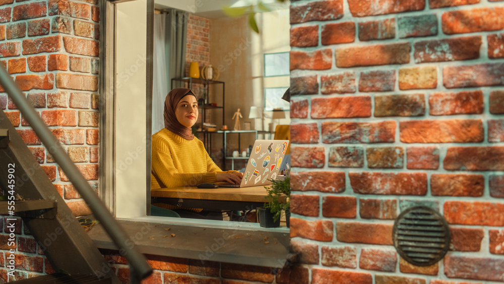 Young Muslim Female Using Smartphone while Sitting on a Window Sill in a Multi-Storey Brick House. Creative Female Smiling and Feeling Happy while Connecting with Family and Friends on Social Media.
