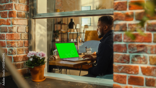 African Man Working on a Laptop Computer with Green Screen Mock Up Display while Sitting at a Table in Stylish Living Room. Business Manager Working Remotely from Home. View from Outside the Window.