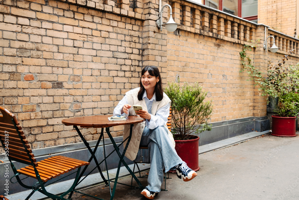 Smiling young asian girl using smartphone while sitting in cafe outdoors