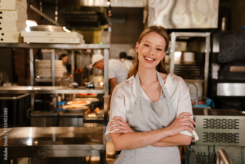 Cheerful woman chef cook standing in kitchen of a restaurant with arms folded