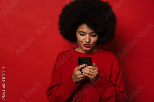Attractive afro woman using smartphone while standing isolated over red studio wall
