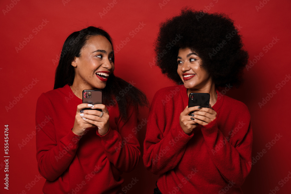 Two smiling multinational women using mobile phones while standing isolated Two smiling multinational women using mobile phones while standing isolated