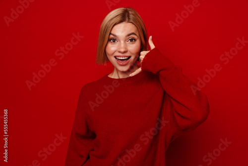 Excited woman showing call gesture and looking at camera isolated over red background