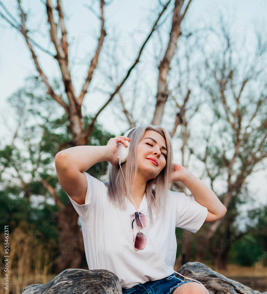 A sweet, young, happy, cheerful, pretty woman sitting outdoors in a park on a beautiful summer day. The girl listens to music with headphones. A sweet, young, happy, cheerful, pretty woman sitting outdoors in a park on a beautiful summer day. The girl listens to music with headphones.