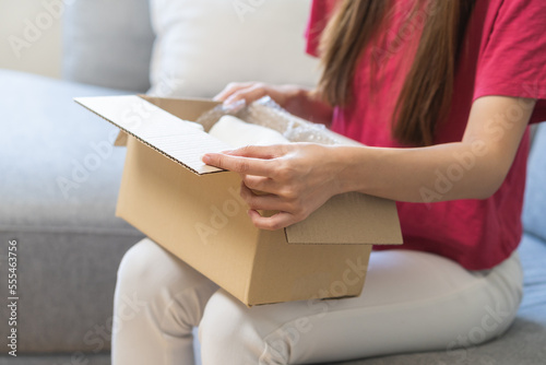 Happy excited, asian young woman, girl customer sitting on sofa at home, open and unpack cardboard box carton parcel after buy fashion clothes, ordering present and shopping online, delivery service.