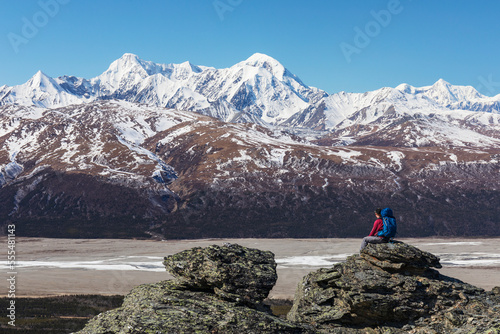 A hiker takes in the view of McGinnis Peak, Mount Moffit, and Mount Hayes in the Eastern Alaska Range while resting on a rock overlooking the Delta River; Alaska, United States of America photo