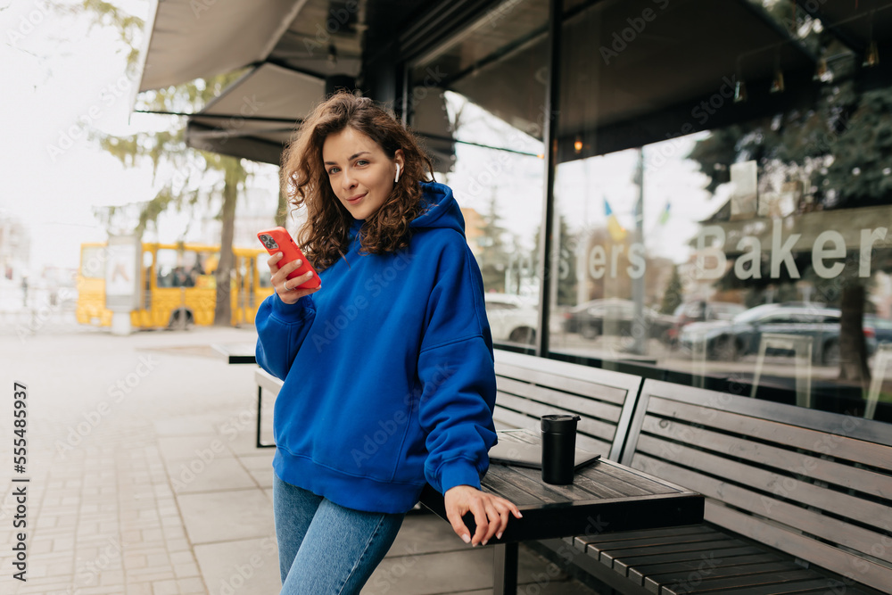 Pretty stylish woman in blue shirt and jeans posing near her house with smartphone. Beautiful lady in trendy outfit walking outside 