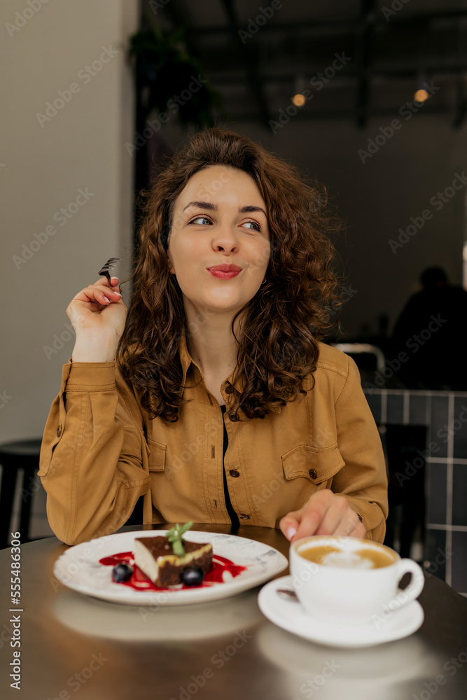 Pretty lovely girl with fluffy girl and nude make up looking aside and enjoying dessert and coffee in city cafe. 