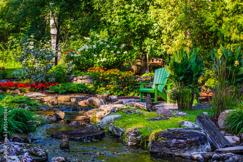 A tranquil stream flows through a garden area with a green adirondack chair; Hudson, Quebec, Canada