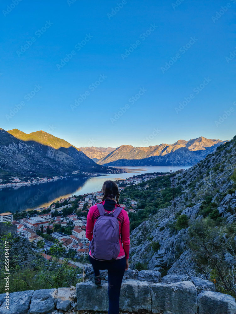 Woman with panoramic view from the Ladder of Kotor on the Kotor bay during sunrise, Adriatic Mediterranean Sea, Montenegro, Balkan Peninsula, Europe. Winding fjord water reflection along coastal towns