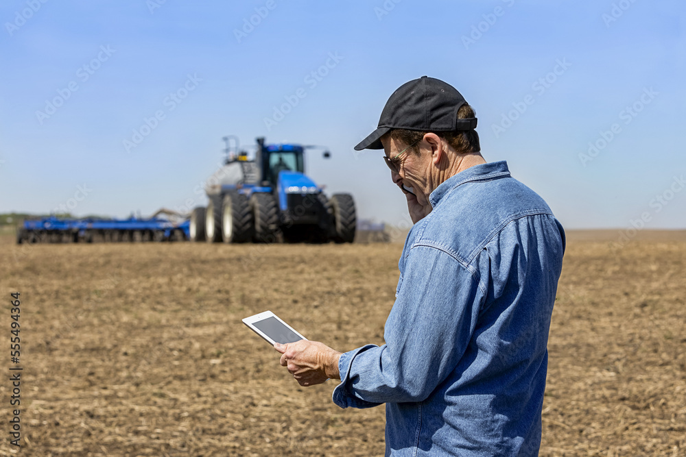 Farmer using a smart phone and tablet while standing on a farm field and watching the tractor and equipment seeding the field; Alberta, Canada Farmer using a smart phone and tablet while standing on a farm field and watching the tractor and equipment seeding the field; Alberta, Canada