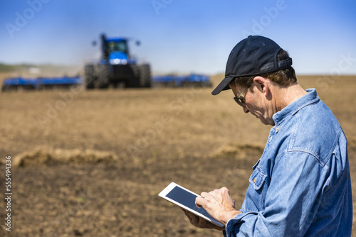 Farmer using a tablet while standing on a farm field and a tractor and equipment seeds the field; Alberta, Canada