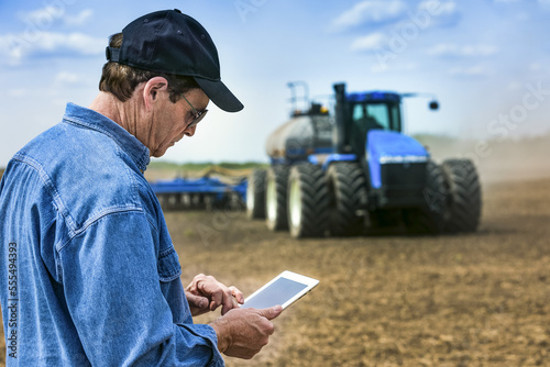 Farmer using a tablet while standing on a farm field and a tractor and equipment seeds the field; Alberta, Canada