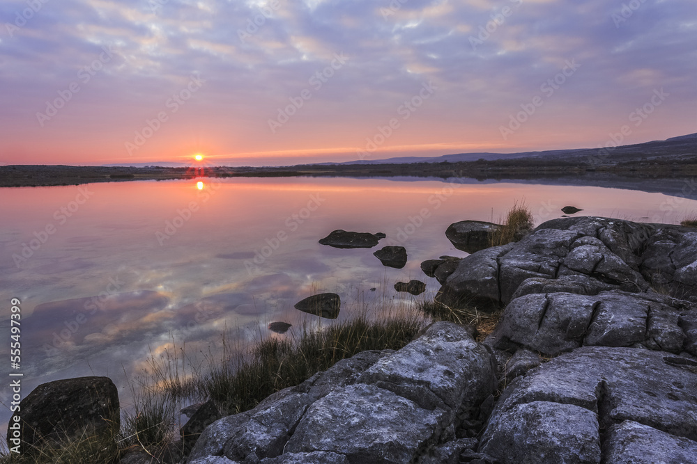 Sun setting over lake with reflections on a cloudy evening with limestone rocks with cracks on the shore in the foreground, Burren National Park; County Clare, Ireland Sun setting over lake with reflections on a cloudy evening with limestone rocks with cracks on the shore in the foreground, Burren National Park; County Clare, Ireland
