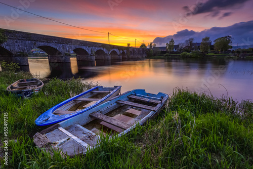 Two small wooden boats on the bank of the Shannon river in front of a stone bridge at sunset in summer; O'Brien's Bridge, County Clare, Ireland photo