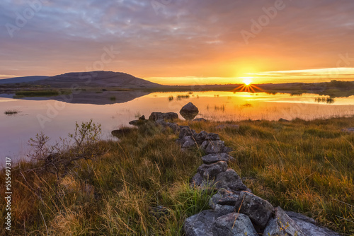 Small stone wall leading to a lake with a mountain and sunrise reflected in the water, Burren National Park; County Clare, Ireland photo