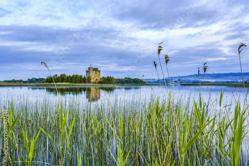 Castlebawn castle on the shore of Lough Derg reflecting in the water on a calm cloudy day in summer with reeds growing in the foreground; Scariff, County Clare, Ireland
