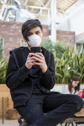 Boy with smart phone wearing protective mask to protect against COVID-19 during the Coronavirus World Pandemic, and his younger sister in the background; Toronto, Ontario, Canada photo