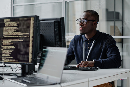 African American young developer in eyeglasses concentrating on his online work on computer sitting at workplace photo