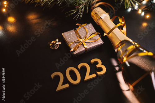 Happy New Year 2023 concept with golden color numbers. Champagne bottle with small gift box and colorful Christmas lights. On a dark background. Top view. Pine tree detail.