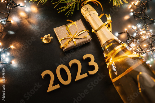 Happy New Year 2023 concept with golden color numbers. Champagne bottle with small gift box and colorful Christmas lights. On a dark background. Top view. Pine tree detail.