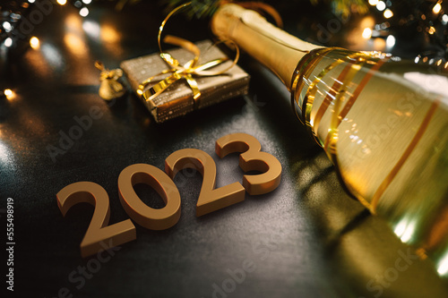 Happy New Year 2023 concept with golden color numbers. Champagne bottle with small gift box and colorful Christmas lights. On a dark background. Top view. Pine tree detail.