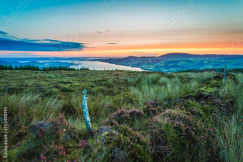 An old wooden fence post on a moor hill overlooking Lough Derg lake in the distance at sunrise; County Clare, Ireland