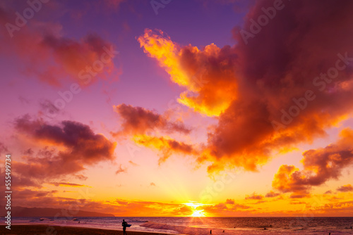 Silhouette of a surfer standing on Kelki Beach at the water's edge with dramatic glowing clouds above at sunset; Oahu, Hawaii, United States of America