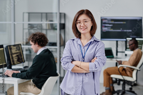 Portrait of Asian young woman smiling at camera standing with her arms crossed in IT office with her colleagues in background