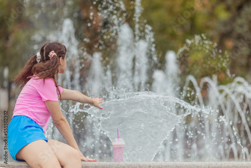 Young happy girl in a candy color clothes pink t-shirt and blue shorts near the city fountain. Cheerful girl having fun in summer vacation. Summer time