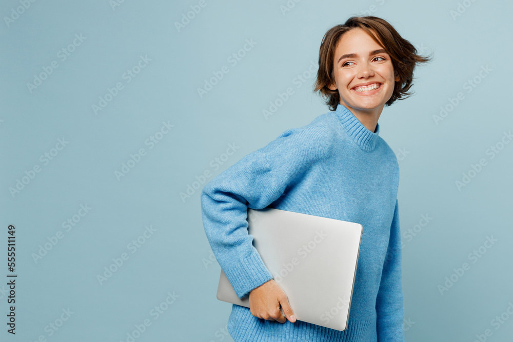 Side view young smiling happy cool caucasian smart IT woman wears knitted sweater hold closed aptop pc computer look aside on workspace area isolated on plain pastel light blue cyan background studio. Side view young smiling happy cool caucasian smart IT woman wears knitted sweater hold closed aptop pc computer look aside on workspace area isolated on plain pastel light blue cyan background studio.