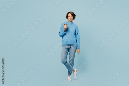 Full body young woman wear knitted sweater look camera hold takeaway delivery craft paper brown cup coffee to go isolated on plain pastel light blue cyan background studio. People lifestyle concept.