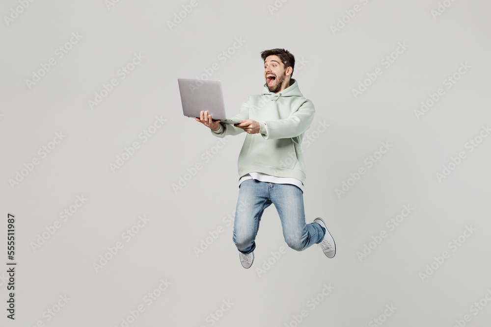 Full body young caucasian happy surprised excited amazed IT man wear mint hoody jump high hold use work on laptop pc computer isolated on plain solid white background studio. People lifestyle concept. Full body young caucasian happy surprised excited amazed IT man wear mint hoody jump high hold use work on laptop pc computer isolated on plain solid white background studio. People lifestyle concept.