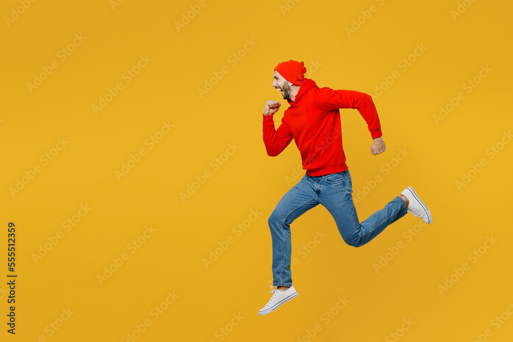 Full body side profile view sporty young caucasian man wearing red hoody hat jump high running fast hurrying in rush isolated on plain yellow color background studio portrait People lifestyle concept