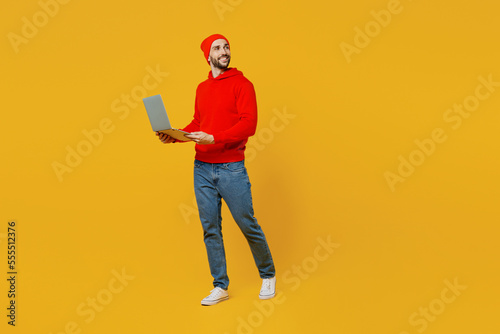 Full body young fun IT man wears red hoody hat look camera hold use work on laptop pc computer look aside on area isolated on plain yellow color background studio portrait. People lifestyle concept.