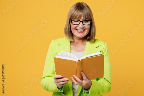 Elderly smiling happy cheerful fun woman 50s years old wearing green jacket white t-shirt reading book novel at library isolated on plain yellow background studio portrait. People lifestyle concept.