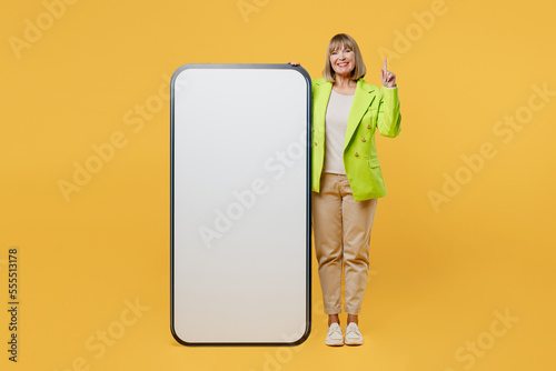 Full body elderly woman 50s years old wear green jacket white t-shirt big huge blank screen mobile cell phone smartphone with area point finger up isolated on plain yellow background studio portrait.