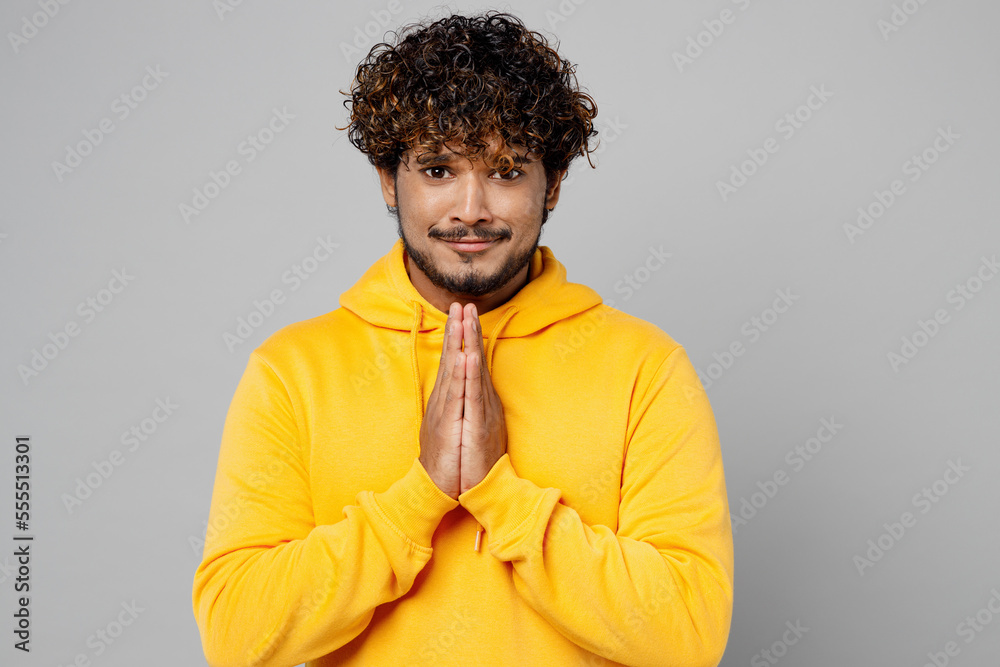 Obraz premium Young asking Indian man 20s he wearing casual yellow hoody holding hands folded in prayer gesture, begging about something isolated on plain grey background studio portrait. People lifestyle portrait.