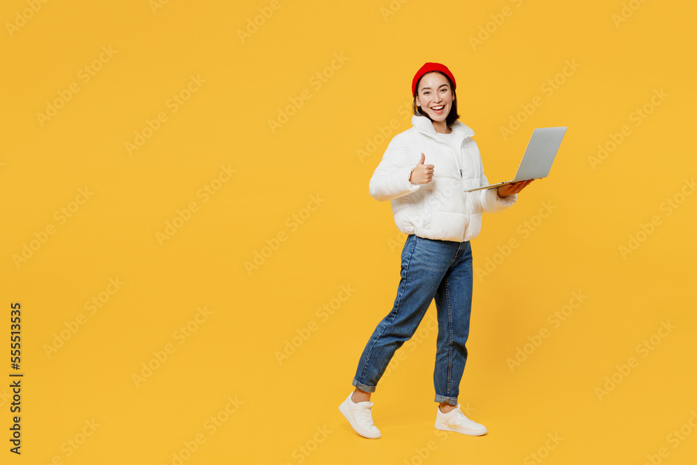 Full body side profile view fun young IT woman of Asian ethnicity wear white padded windbreaker jacket red hat hold use work on laptop pc computer isolated on plain yellow background studio portrait. Full body side profile view fun young IT woman of Asian ethnicity wear white padded windbreaker jacket red hat hold use work on laptop pc computer isolated on plain yellow background studio portrait.