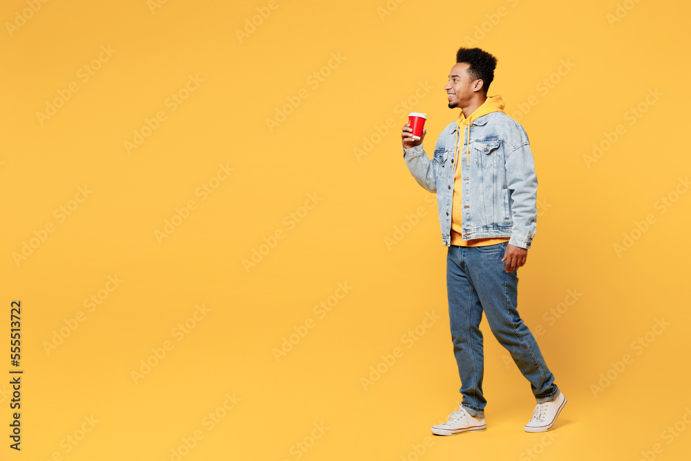 Fototapeta premium Full body side view young man of African American ethnicity wear denim jacket hoody hold takeaway delivery craft paper brown cup coffee to go walk isolated on plain yellow background studio portrait.