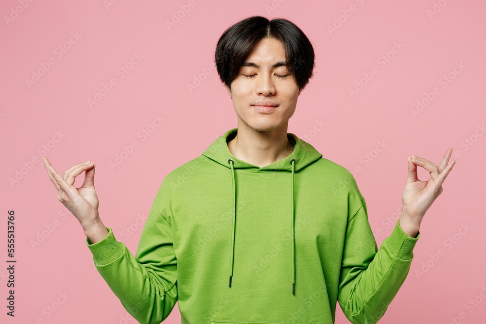 Young man of Asian ethnicity wear green hoody hold spread hands in yoga om aum gesture relax meditate try to calm down isolated on plain pastel light pink background studio People lifestyle concept