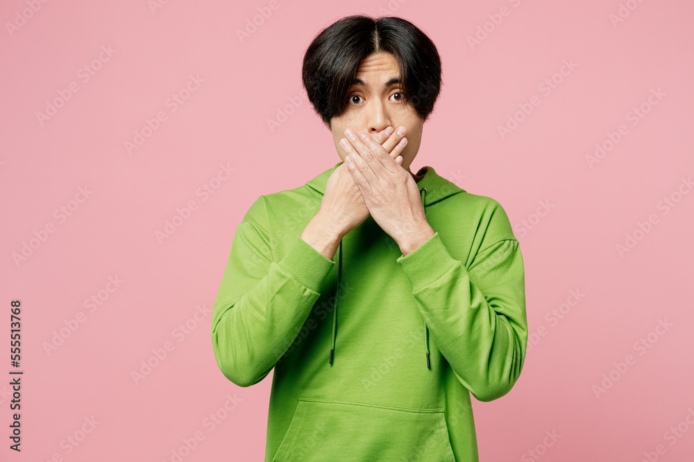 Young scared sad fearfur shocked man of Asian ethnicity he wears green hoody look camera cover mouth with hand isolated on plain pastel light pink background studio portrait. People lifestyle concept. Young scared sad fearfur shocked man of Asian ethnicity he wears green hoody look camera cover mouth with hand isolated on plain pastel light pink background studio portrait. People lifestyle concept.