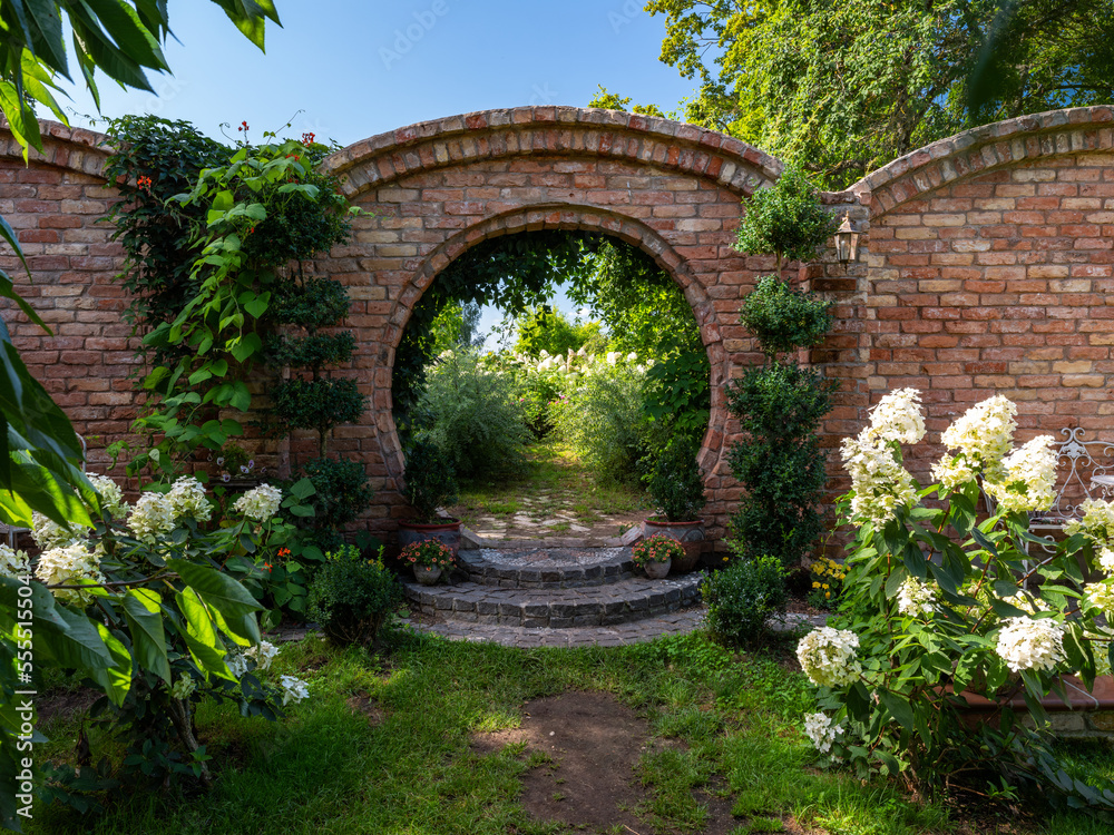 Beautiful summer garden at sunny day. Flowers, plants, bushes. Arch in brick wall