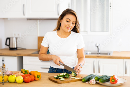 Positive young latin american girl standing in cozy kitchen interior at home, slicing fresh vegetables for salad