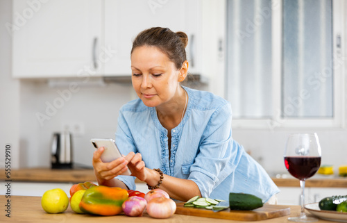 Portrait of young adult woman browsing on her phone while cooking at home kitchen