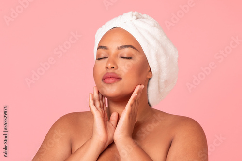 Portrait of body positive black woman with smooth skin touching her face while posing over pink studio background