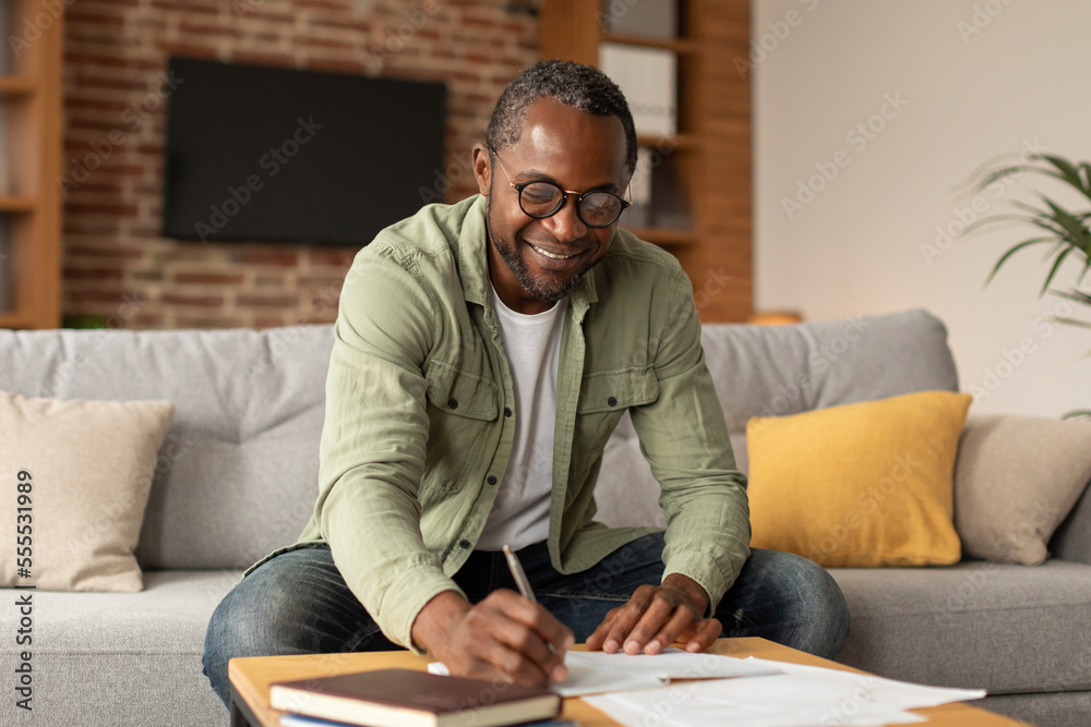 Fototapeta premium Cheerful adult black businessman in glasses makes notes, studies and works in living room interior, copy space